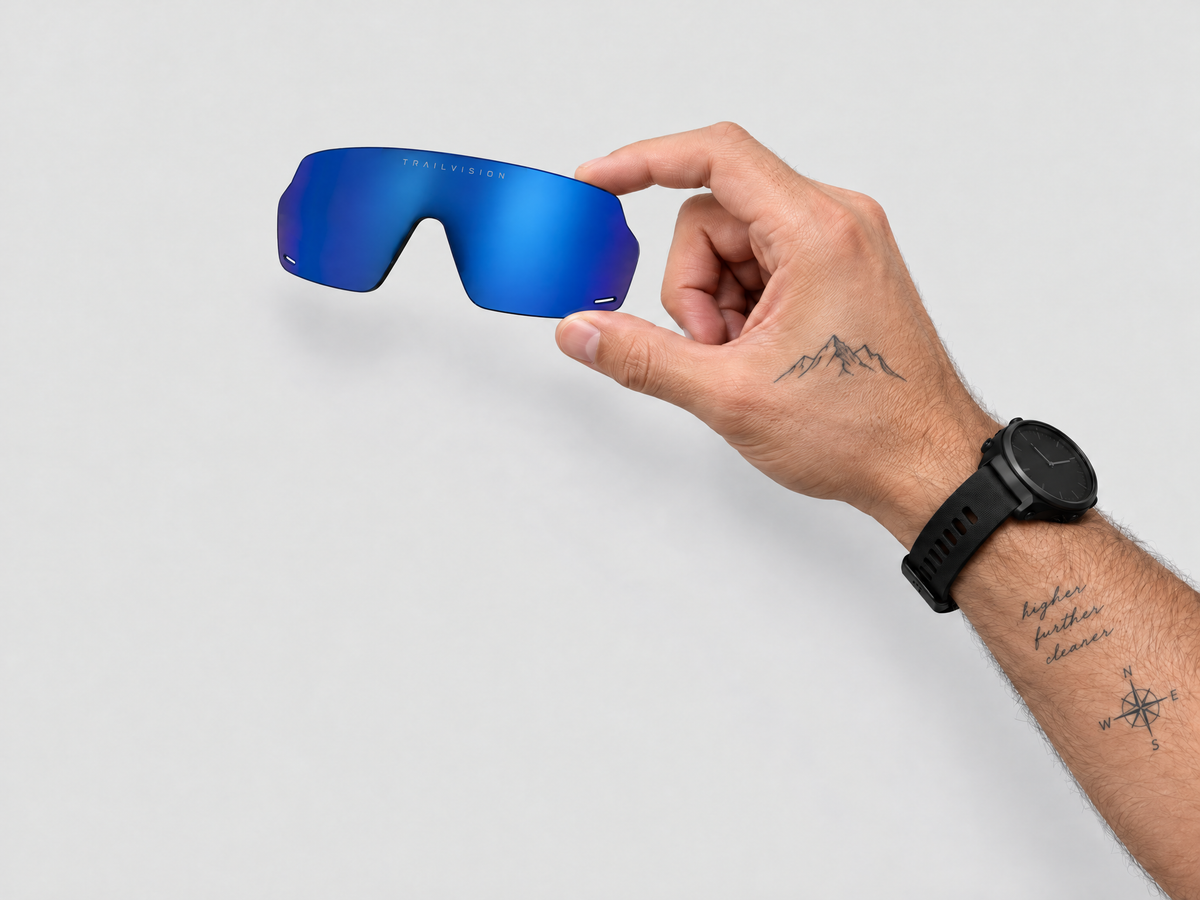 Hand holding a blue mirrored Trailvision replacement lens with laser-engraved TRAILVISION on a light gray background